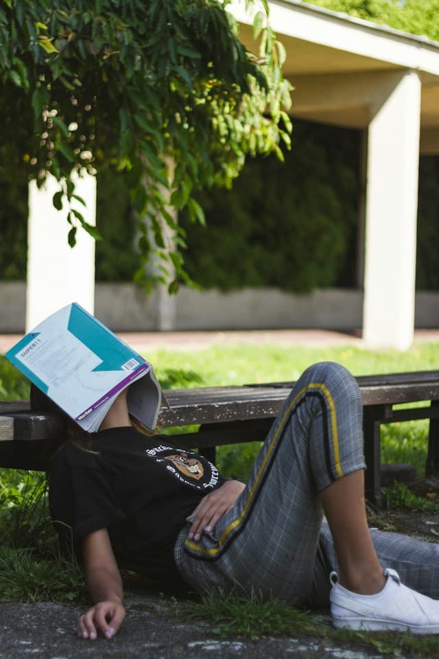 A person relaxing outdoors with an open textbook covering their face.