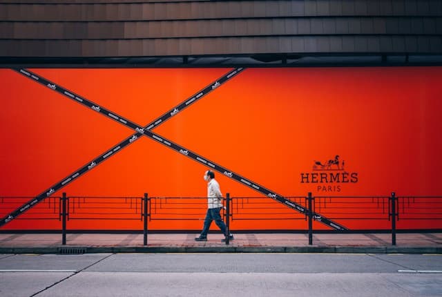 A pedestrian walks in front of an enormous vivid orange Hermès Paris storefront display crossed by black branded ribbons.