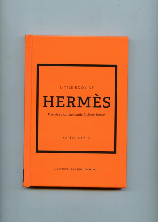 An orange hardcover copy of Little Book of Hermès by Karen Homer photographed against a pale grey background.